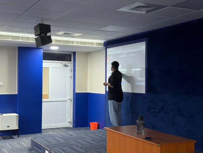 A person standing in a blue and white room presenting in front of a projection screen, with a speaker mounted on the ceiling.