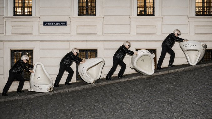 A staged photograph showing four identical figures of an older man in black clothing lined up against a classical building wall with barred windows, each pushing or tilting a large white ceramic object on a cobblestone street.