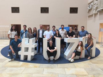 A group photo of around 15 people posing together in front of large '#UZH' letters in what appears to be a university or institutional lobby.