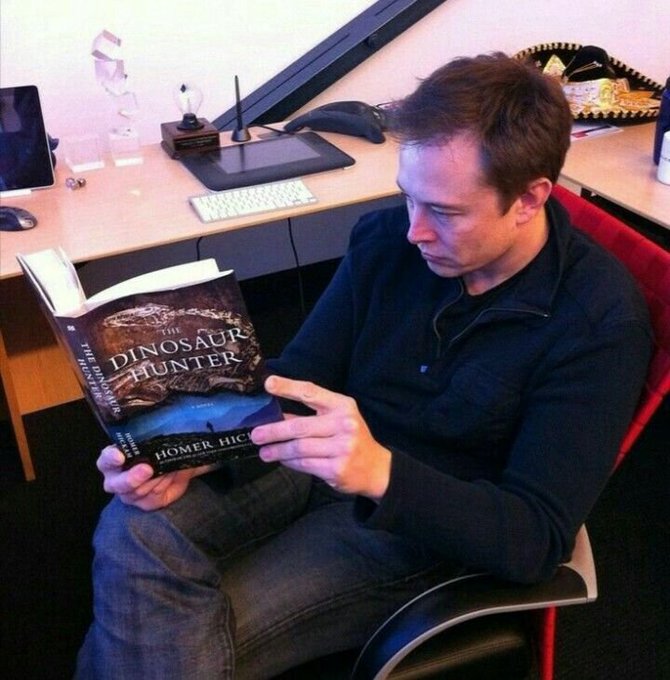 A man sits in an office chair reading a book titled 'The Dinosaur Hunter' by Homer Hickam, with a desk and computer monitor visible in the background.