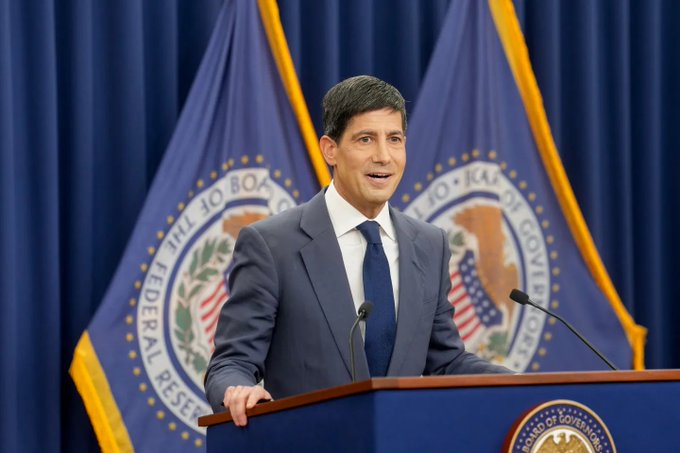 A man in a dark suit stands at a podium with Federal Reserve Board of Governors seals and flags in the background, speaking at a press briefing. The image relates to the FOMC minutes announcement, as the setting depicts an official Federal Reserve speaker, fitting the post's discussion of the upcoming minutes release under Fed leadership.