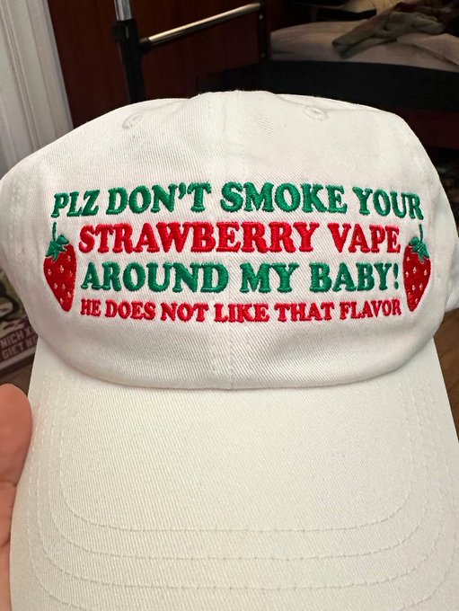 A hand holds up a white baseball cap with embroidered text reading 'PLZ DON'T SMOKE YOUR STRAWBERRY VAPE AROUND MY BABY! HE DOES NOT LIKE THAT FLAVOR' flanked by two strawberries.