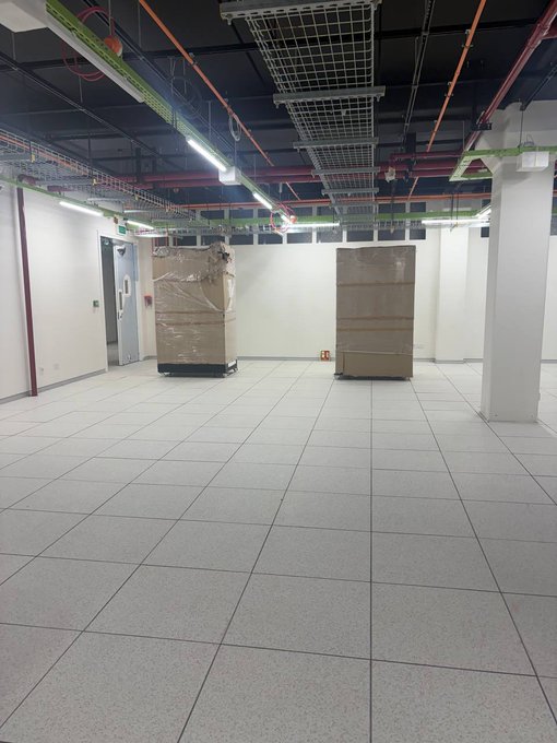 An empty room with a tiled floor, white walls, exposed ceiling wiring, and two large objects wrapped in plastic.
