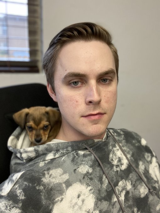 A young man takes a selfie while cradling a small brown and black puppy against his chest, looking calmly at the camera. The relaxed, cozy scene complements the giveaway winner announcement by suggesting a personal moment shared with the recipient.