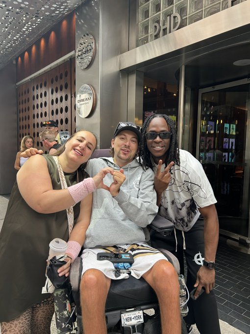 Three people posing together outside a venue, with the person in the middle making a heart shape with their hands.