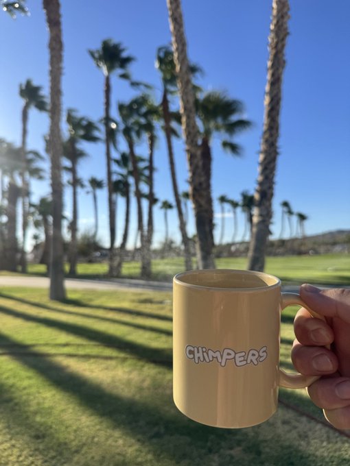 A hand holds a tan ceramic mug with 'CHIMPERS' written on it in front of a scenic background of tall palm trees and blue sky.