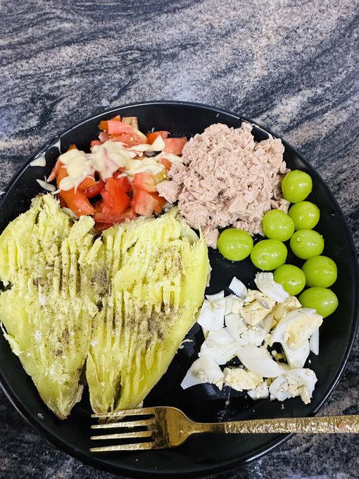 A heart-shaped avocado is served on a black plate alongside tuna, tomatoes, green grapes, and egg, with a gold fork — a wholesome, love-themed meal signaling a cozy night in.