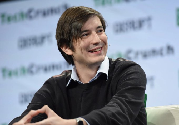 A young man with brown hair smiles while speaking at what appears to be the TechCrunch Disrupt event, wearing a dark sweater over a collared shirt.