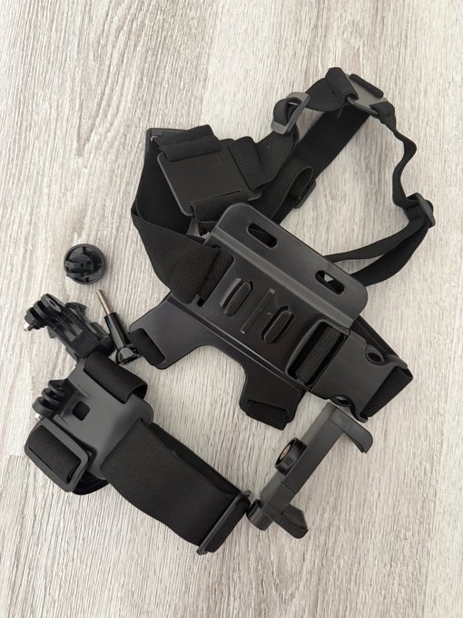 The image features a black strap with a camera attached, likely used for attaching cameras to body armor or other protective gear. There are two additional items present in the scene - one of them being a black phone case. The main focus of the image is on the strap and its connection to the camera, while the background appears to be a wooden surface.