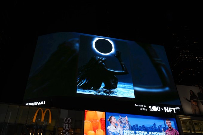 A nighttime photograph of a large digital billboard in Times Square displaying an NFT artwork of a figure under an eclipse, with text reading 'Curated by Stills 100 NFT', positioned above a McDonald's sign.
