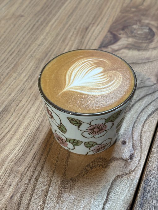 A ceramic cup decorated with pink cherry blossoms, filled with a latte featuring intricate leaf-patterned latte art, resting on a textured wooden table.