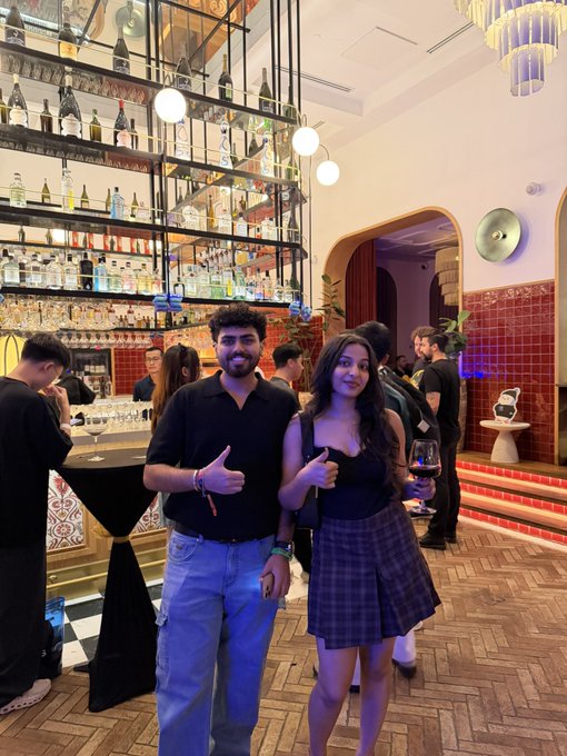 A man and woman pose for a photo at a stylish bar with shelves of liquor bottles in the background, both giving thumbs up while the woman holds a glass of red wine. The setting reflects a social meetup vibe, fitting the caption's reference to meeting a notable person in a lively, upscale venue.