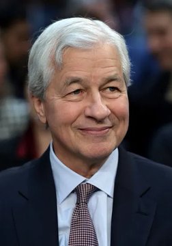 A close-up photograph of an older man with silver-gray hair wearing a dark suit, white shirt, and patterned tie, smiling slightly at the camera.