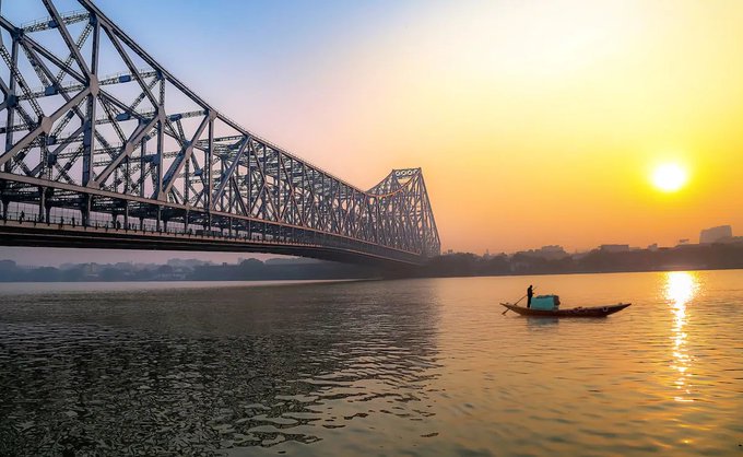 A sunset view of the Howrah Bridge in Kolkata, India, with a small boat floating on the Hooghly River in the foreground.