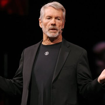 A man with gray hair and a beard, wearing a black blazer over a black t-shirt with a circular pendant, speaks on a stage.
