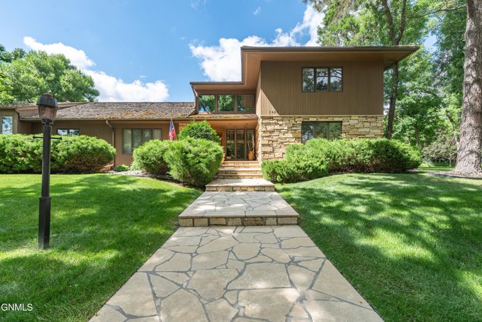 A real estate listing photo (GNMLS watermark) showing a mid-century modern style home with wood and stone exterior, a flagstone walkway, manicured lawn, and a Texas flag near the entrance.
