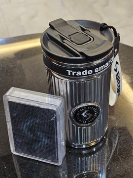 A black 'Trade Smart' trash can-shaped container next to a small clear case with a mesh-like design, sitting on a dark countertop.
