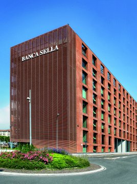 The image shows a large, red brick building with the name "BANCA SELLA" displayed prominently on its side. The building also has a sign that reads "BITCOIN & CRYPTO SERVICES IS EMBRACING BTC." This suggests that the bank is now offering Bitcoin and crypto services to its customers.

The scene takes place in front of a street, with a parking lot visible behind the building. There are several cars parked along the street, indicating that it might be a busy area or a popular location for visitors.