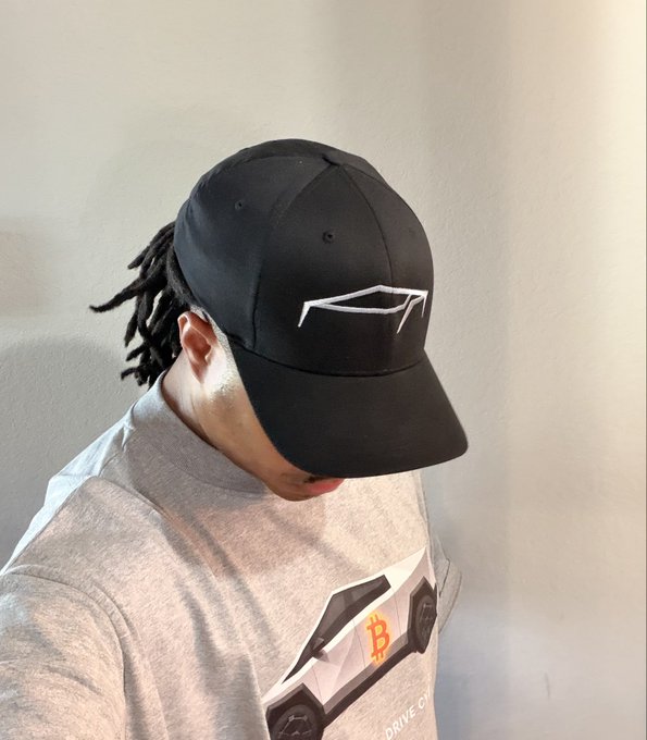 A person wearing a black baseball cap with a white geometric logo and a gray t-shirt featuring a Bitcoin graphic and 'DRIVE' text.