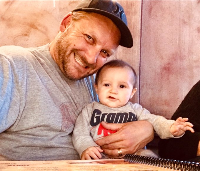 A smiling man wearing a baseball cap holds a baby dressed in a grey shirt that says 'Grandpa' in blue and red letters.