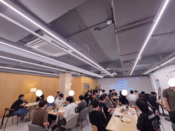 A group of people sits at long tables in a modern office space, eating together in front of a projection that reads 'Renoise KR Community'.