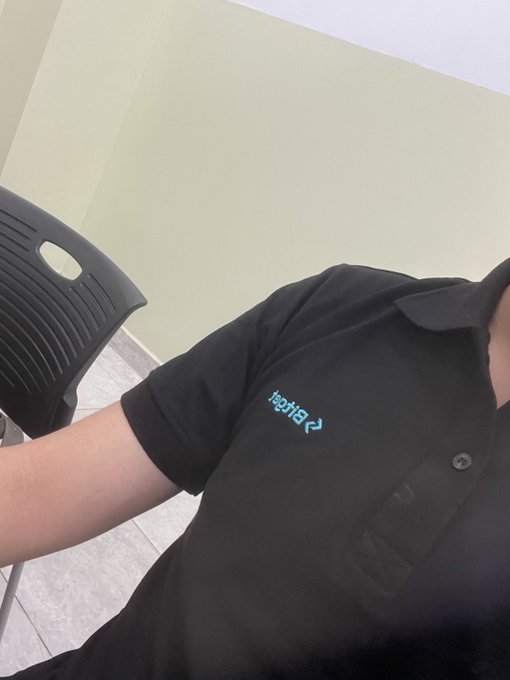 A person wearing a black Bitget-branded polo shirt sits next to a black plastic chair in a room with light green walls.