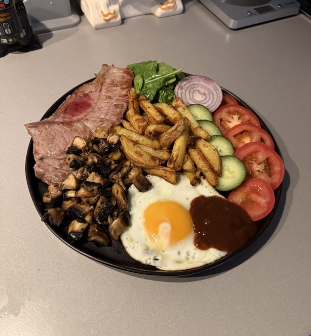 A black plate holds a hearty meal featuring sliced steak, sautéed mushrooms, french fries, a fried egg, BBQ sauce, and garnishes of cucumber, tomato, red onion, and spinach.
