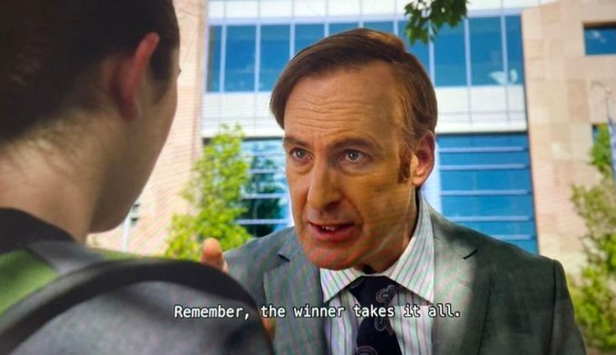 A suited man speaks intently to another person outdoors; the subtitle reads, “Remember, the winner takes it all.”