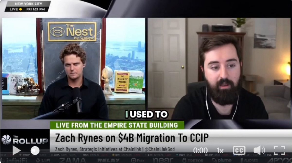 The image shows a man with a beard sitting at a desk, wearing a black shirt. He has a microphone in front of him, suggesting that he might be hosting a podcast or recording an interview. The background features a window overlooking a cityscape, giving the impression that they are in an urban setting. There is also a book visible on the desk next to the man with the microphone.