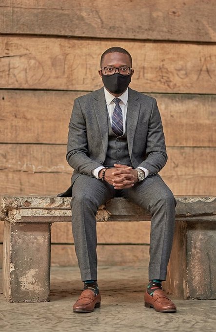 A sharply dressed man wearing glasses and a black face mask sits on a weathered stone bench against a wooden plank wall, wearing a grey three-piece suit, striped tie, and brown shoes with colorful socks.