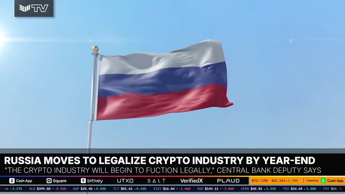 A Russian flag waves against a blue sky above a news ticker reading "RUSSIA MOVES TO LEGALIZE CRYPTO INDUSTRY BY YEAR-END" and "*THE CRYPTO INDUSTRY WILL BEGIN TO FUCTION LEGALLY*,* CENTRAL BANK DEPUTY SAYS," illustrating Russia's reported push to formally regulate Bitcoin and crypto activity by the end of 2026.
