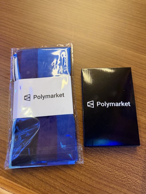Two Polymarket promotional items sit on a wooden surface: a blue packaged item and a black card holder, both displaying the Polymarket logo.