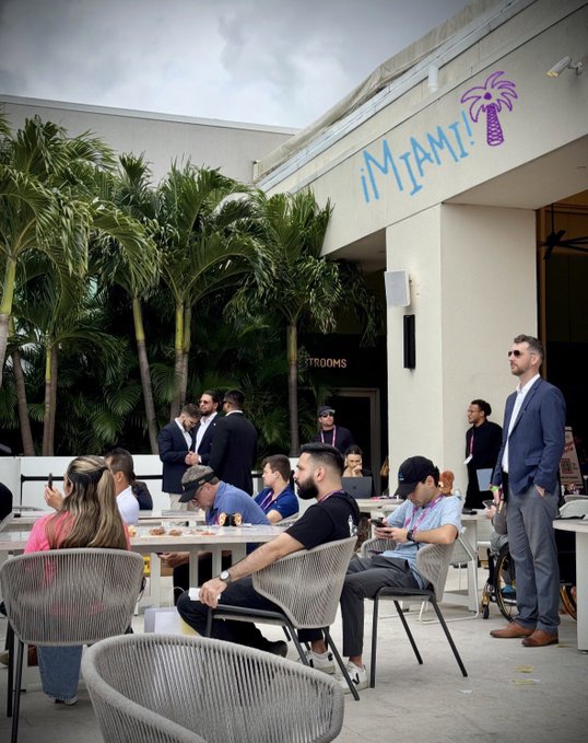 People gather at an outdoor venue featuring a large "MIAMI!" sign with a palm tree logo, dining at tables under cloudy skies with security and staff in attendance. The "DDMIA" caption suggests this is an event or meetup taking place in Miami.
