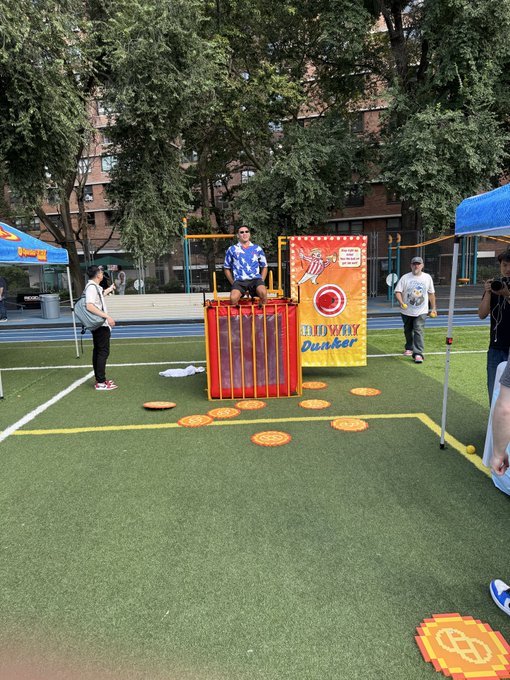 An outdoor event featuring a Dunkin'-branded "Dunk Way Dunker" dunk tank game, where a man in a blue patterned shirt is seated on the dunk seat above a field marked with paddle targets.