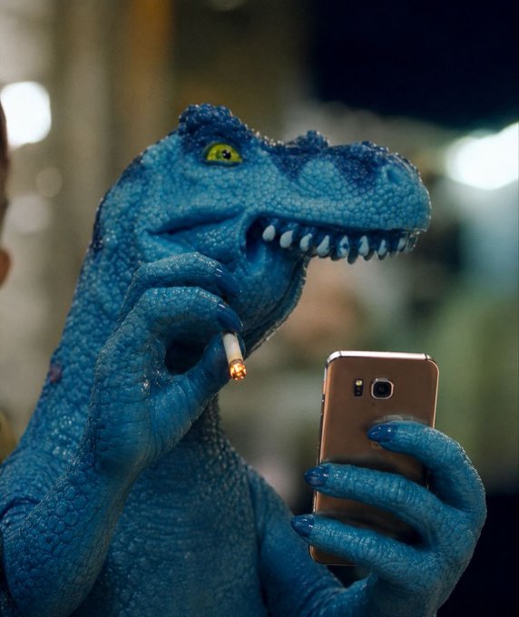 A blue, scaly reptilian figure reminiscent of a raptor smokes a cigarette while scrolling through a rose-gold smartphone, suggesting a humorous, irreverent meme about "aping" into crypto trades.

The image plays on the post's tone by visually personifying reckless trading behavior—combining a prehistoric predator (impulsive risk-taking) with the casual habit of checking a phone (scanning markets)—as a witty nod to the user's CoinGecko ID.
