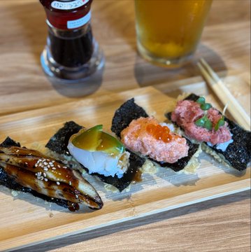A wooden sushi platter features an assortment of gunkan-maki nigiri topped with eel, avocado, salmon roe (ikura), and spicy tuna, served alongside a glass of beer and a dark bottle, pairing a casual meal with the social media post's playful reference to "bullposting" from a pub or restaurant setting.