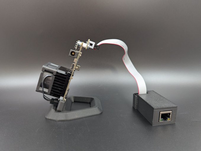 A custom electronics project featuring a small camera module with a heat sink mounted on a 3D-printed stand, connected via a ribbon cable to an Ethernet adapter housed in a 3D-printed case.