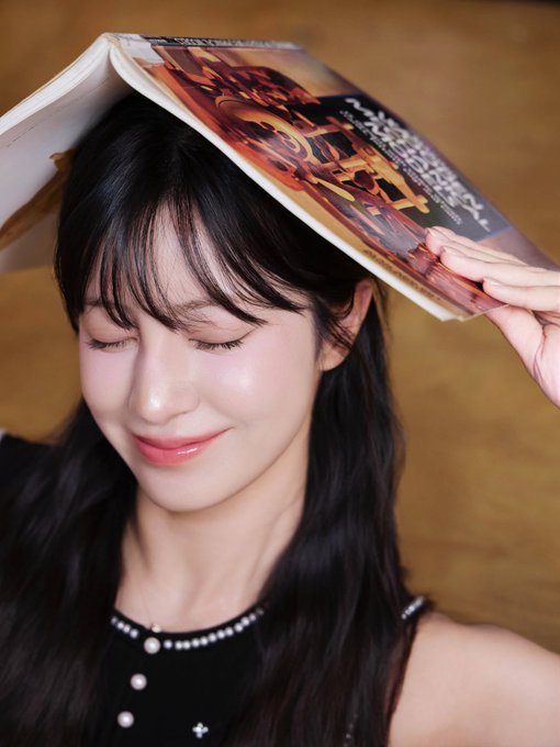 A woman with long dark hair smiles with her eyes closed while balancing a magazine on her head, wearing a black top with pearl details.