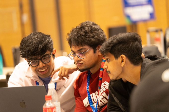 Three young men collaborating and smiling while looking at an Apple MacBook laptop at what appears to be a hackathon or tech event, with one wearing a red "University" shirt.