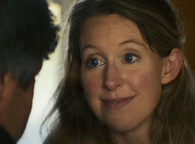A close-up cinematic shot of a woman with long wavy blonde hair and blue eyes looking with wide-eyed surprise at a man whose back is to the camera.