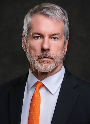 A portrait of a distinguished man with gray hair and a beard, wearing a black suit, white shirt, and orange tie, against a dark background.

The image likely depicts Michael Saylor, the MicroStrategy chairman known for his outspoken support and ongoing large-scale purchases of Bitcoin for his company's treasury.