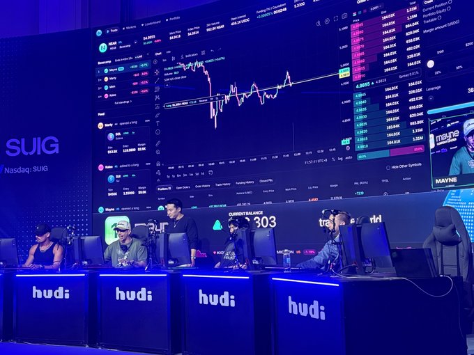 Photo of an esports tournament setup with multiple players seated at gaming stations under blue lighting, with large screens displaying cryptocurrency trading charts including SUIG token and 'hudi' branding on the desks.