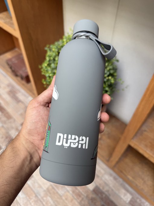 A hand holds a matte gray insulated water bottle featuring a Dubai skyline design and the word "DUBAI" in both English and Arabic, set against a background of wooden furniture and a small plant.