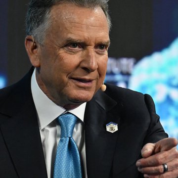 A close-up photograph of Robert F. Kennedy Jr., wearing a dark suit with a blue tie and a hexagonal pin on his lapel, looking off-camera with a slight smile.