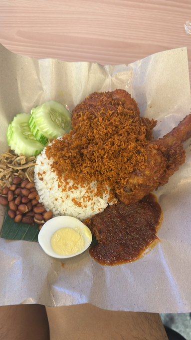 A photograph of a meal of nasi lemak served on paper, featuring fried chicken topped with sambal, rice, peanuts, cucumber, anchovies, and a halved boiled egg.