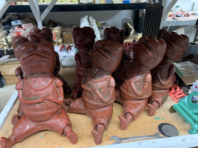 Several reddish-brown sculptures of chubby frogs wearing hoodies are arranged on a workbench in what appears to be an artist's studio.