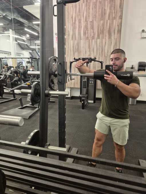 A man in an olive green t-shirt and cream shorts takes a mirror selfie at a gym, standing near a squat rack with weight plates loaded on it.