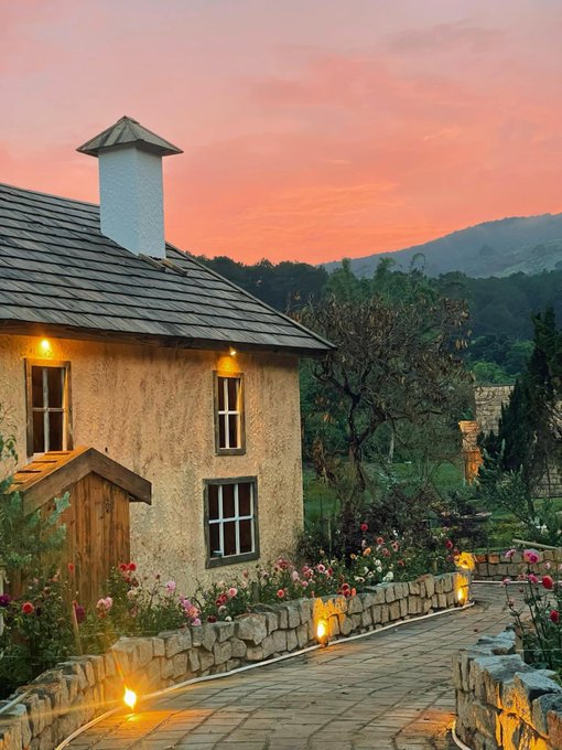 A charming stone cottage with a lit pathway at sunset, surrounded by flowers and rolling hills.