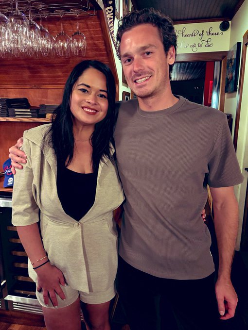 A woman with long black hair in a beige blazer dress stands next to a tall man with curly hair wearing a brown t-shirt in what appears to be a restaurant or bar setting.
