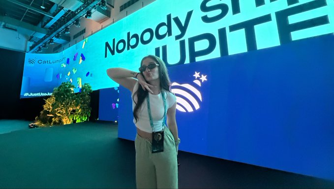 A woman with long dark hair wearing sunglasses, a white crop top, and beige pants poses in front of a large screen at what appears to be a tech event, with the text 'Nobody SH JUPITE' and 'CatLumi' partially visible.